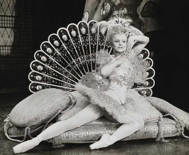 A ballerina in costume reclines on cushions with a stylized peacock tail fanned out behind her.