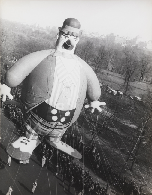 Um grande balão de palhaço flutua pelas ruas da cidade, com uma multidão de espectadores abaixo.