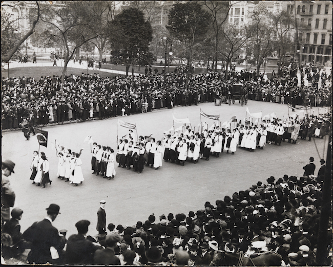 Black and white photo featuring a parade of women with signs heading down an empty street flanked with crowds of spectators.