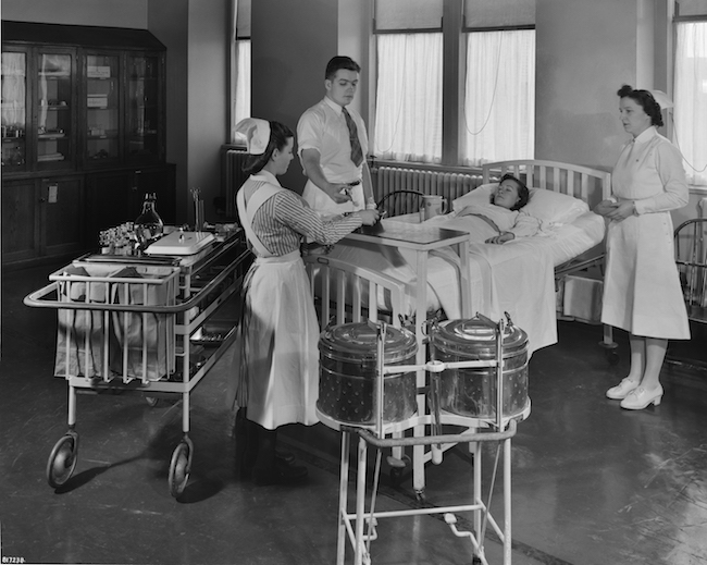 Uma fotografia em preto e branco de três enfermeiras em pé ao redor de um paciente em uma cama de hospital.