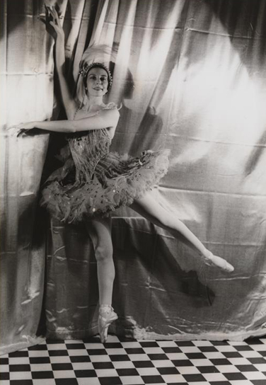 A ballerina, in costume, stands en pointe in front of a curtain on a checkerboard floor.