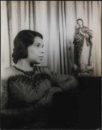 Black and white portrait of Marian Anderson in front of a curtain. Her arms rest on a dark surface, with a statue of a religious figure next to her left elbow.