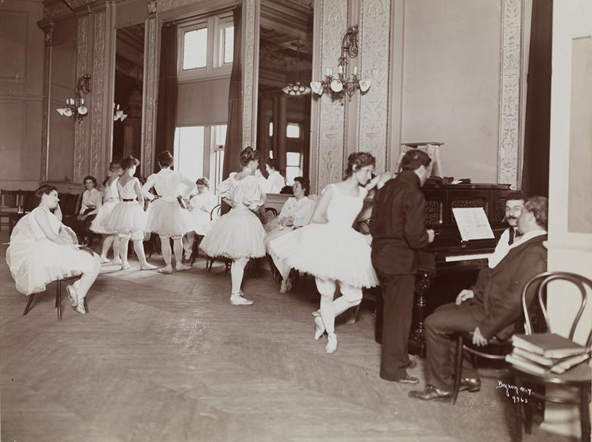 A group of ballerinas are gathered in a large room. In the background, some stand, and others sit. In the lower right, one ballerina leans against a piano with three gentleman, one standing, and two sitting at the instrument. 