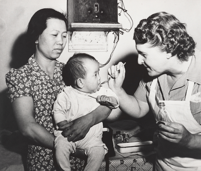 Photographie en noir et blanc d'une infirmière nourrissant un bébé d'origine asiatique à la cuillère, tenue par sa mère.