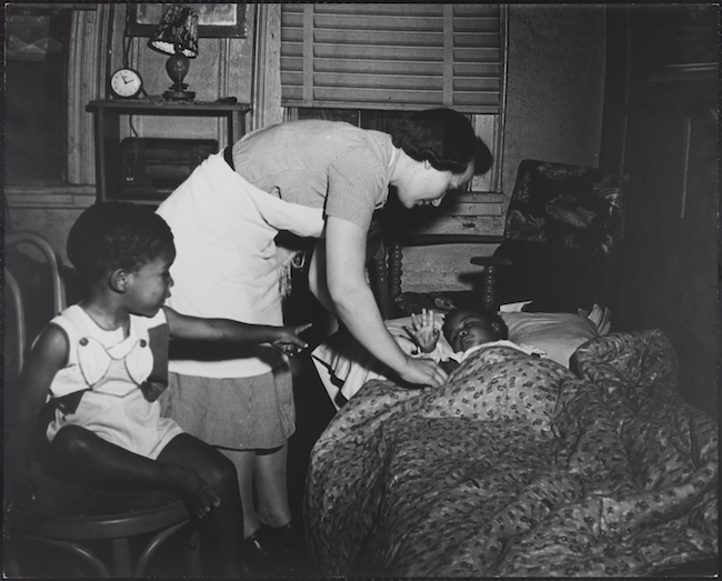 A nurse tucks a small child into bed, while another, seated in a chair, looks on.