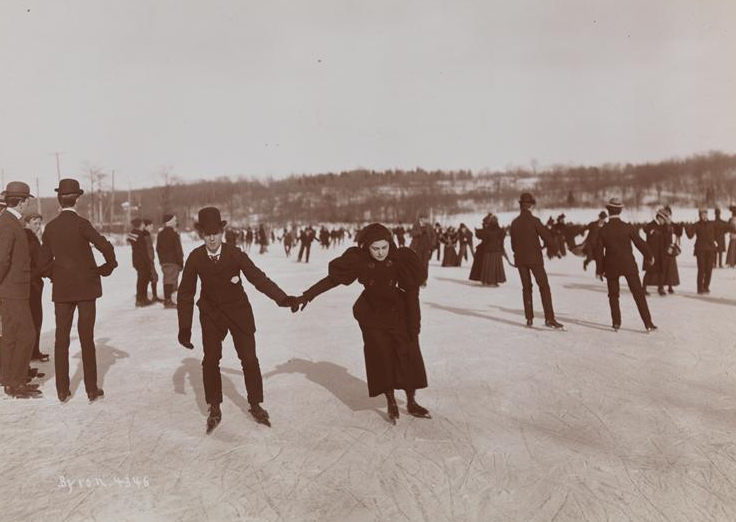 A man and women hold hands while ice skating among a group of people outdoors.