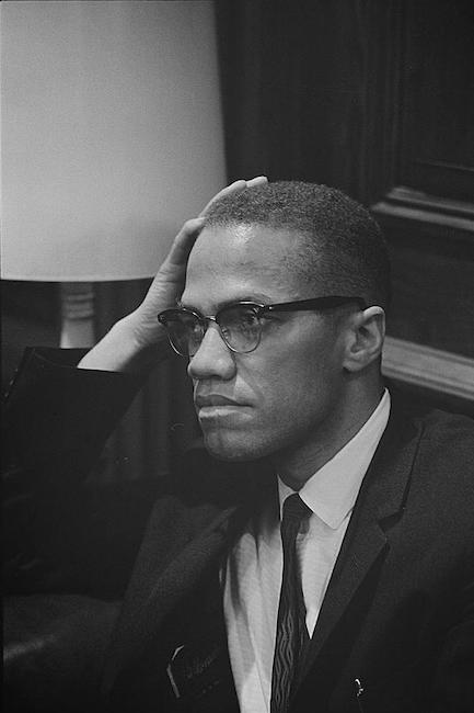 Photograph shows Malcolm X, sitting in a room with his hand on his head.