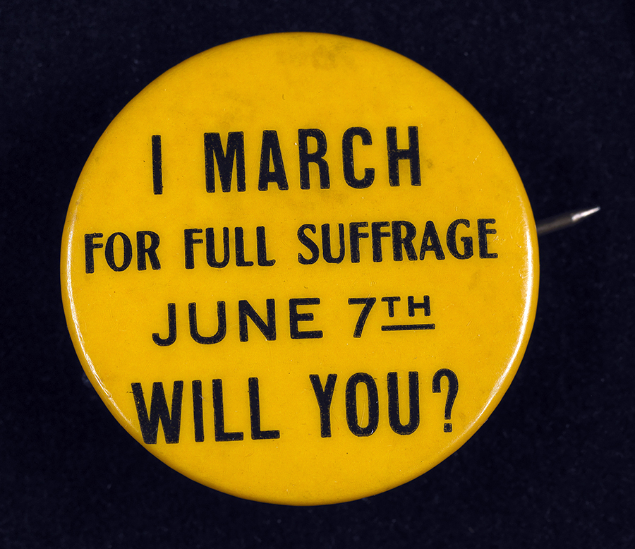 A small gold button supporting voting rights for women from the early 20th century reads: I March for Full Suffrage June 7th. Will You? 