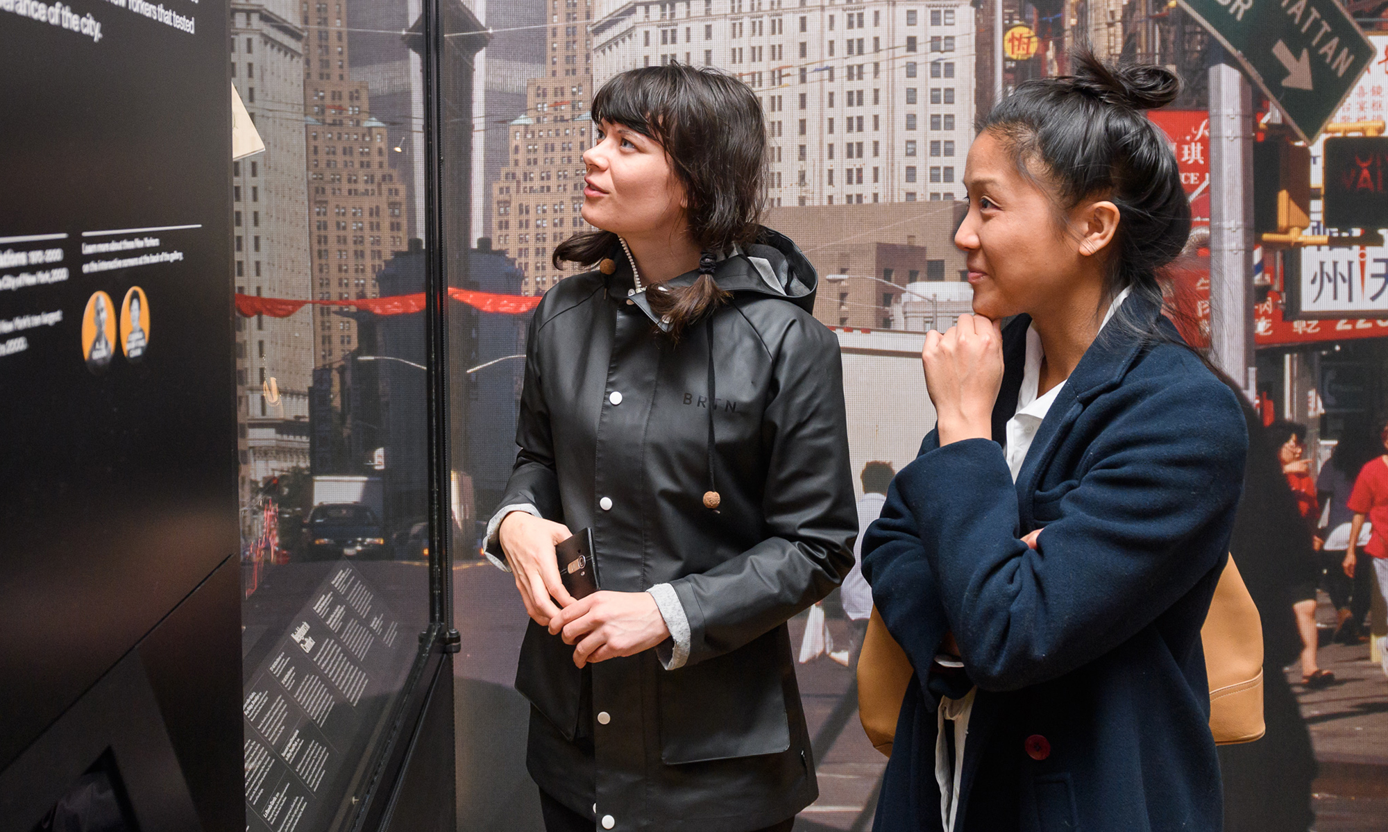 Two people in front of a photograph of New York City, looking at exhibition objects