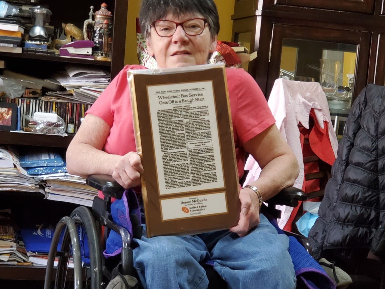 Une femme à lunettes dans un fauteuil roulant brandit un article de presse encadré.