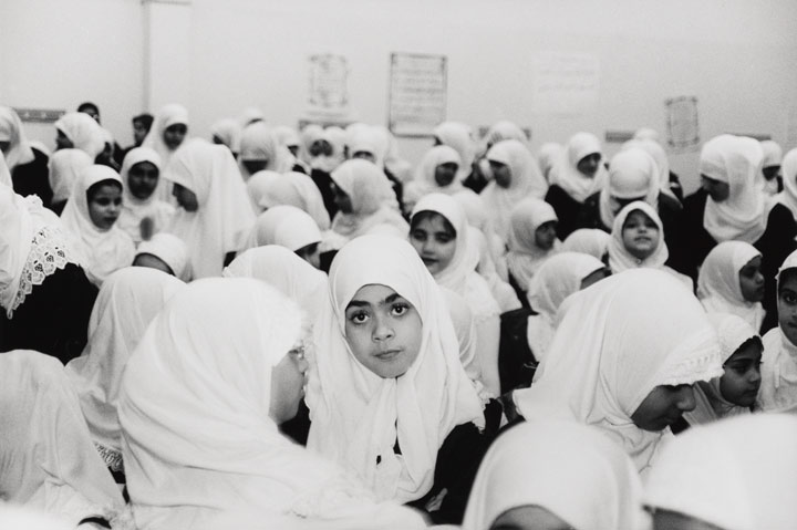 A group of girls wearing hijabs stand in a schoolyard, one girl in the middle stares into the camera