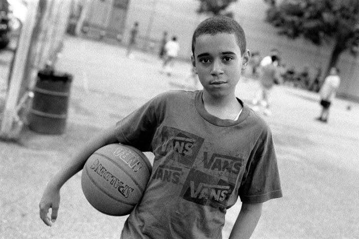 一个男孩拿着篮球时凝视着镜头。 在他身后，其他孩子在球场上打篮球