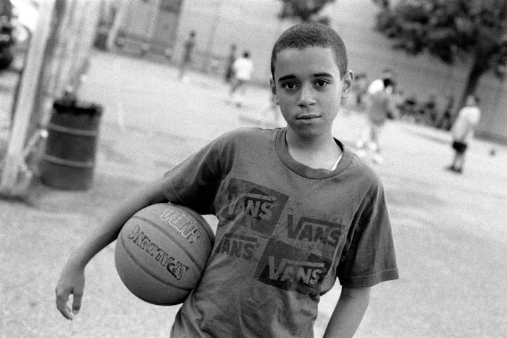 Un niño mira a la cámara mientras sostiene una pelota de baloncesto. Detrás de él, otros niños juegan baloncesto en una cancha