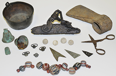 Photograph of dark metal pot, stone arrow heads, colorful string of painted beads, stone carvings, and other objects together.