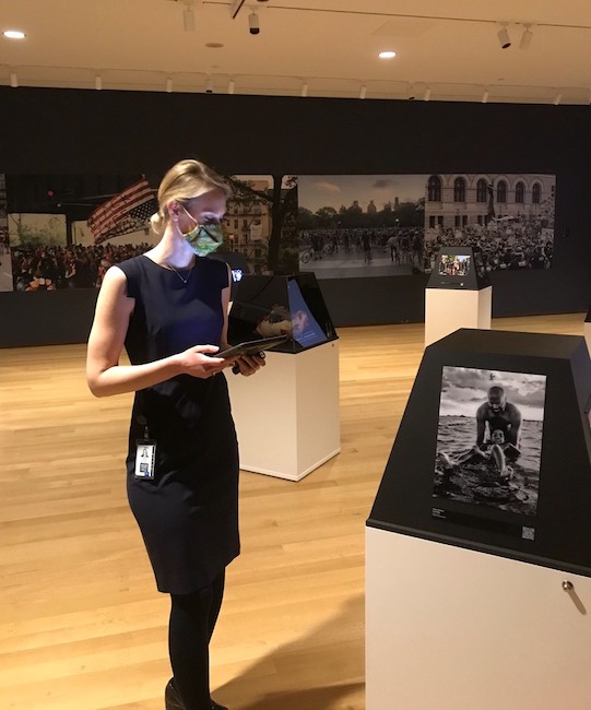 A woman wearing a face mask stands in the gallery of the exhibition "New York Responds," viewing one of the works on view on the plinths set up throughout the space.