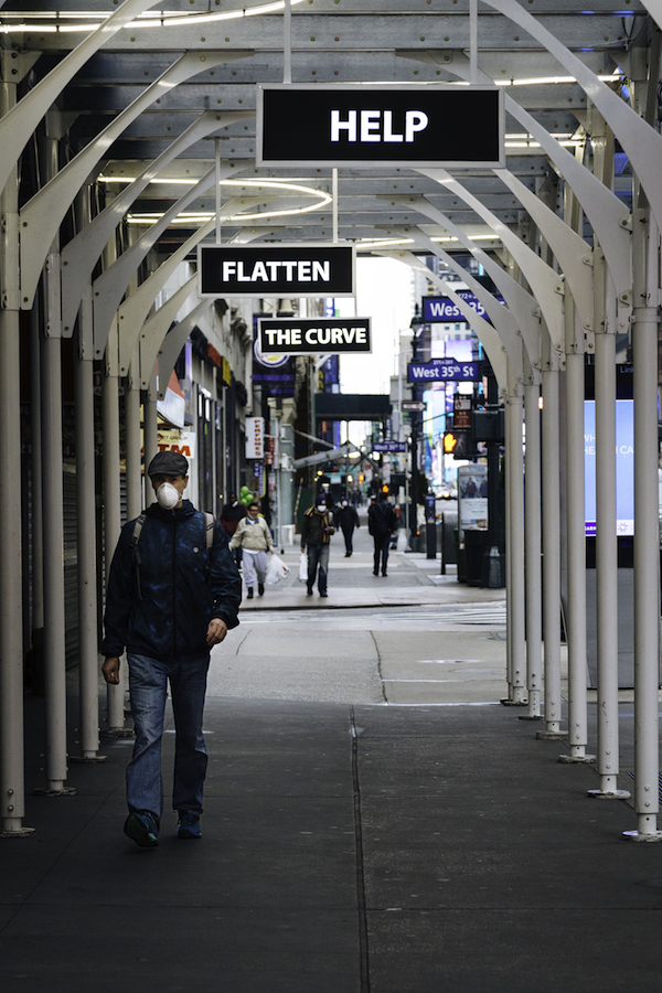 A man walks through a covered section of the side walk. Three signs hang from the top with the words "HELP" "FLATTEN" "THE CURVE"