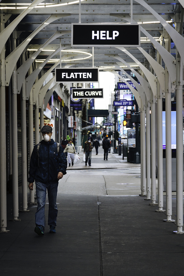 男が歩道の覆われた部分を歩きます。 「HELP」「FLATTEN」「THECURVE」の文字が上からXNUMXつの看板にぶら下がっています