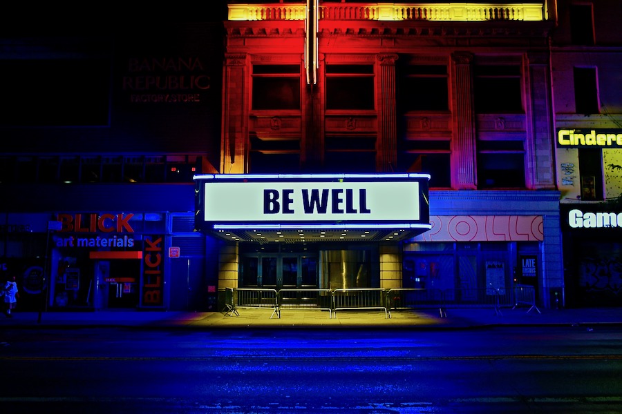 The words “Be Well” are illuminated on the marquee of the Apollo Theater in Harlem. Blue and red lights are projected on the building.