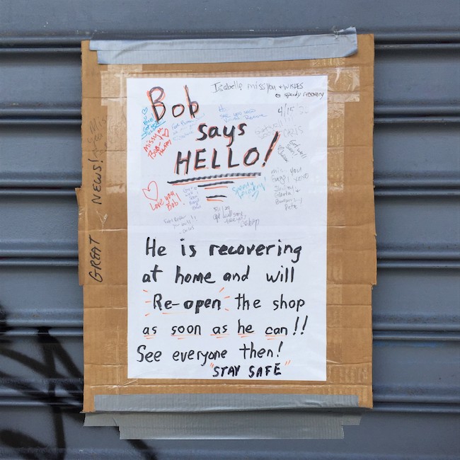 Sign, written on white paper taped on to cardboard, is duct taped to the metal grate on a closed storefront. Sign reads "Bob says Hello! He is recovering at home and will Re-open the shop as soon as he can!! See everyone then ! STAY SAFE. Small, personal messages from multiple people appear in the space around these words.