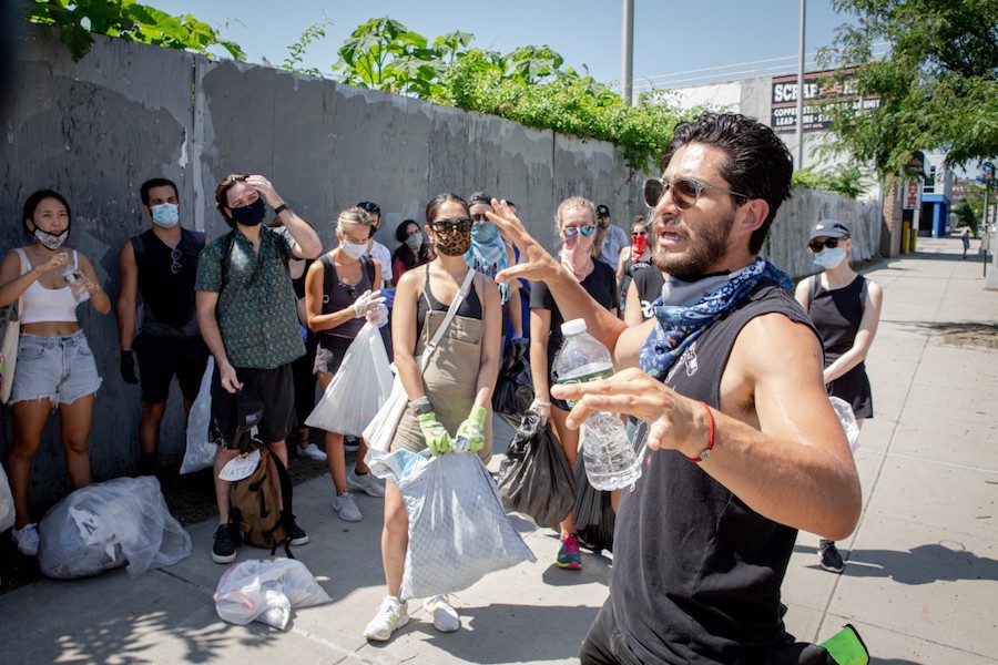 A man holding a water bottle in one hand addresses a crowd of people with bags and masks standing in front of a wooden fence.
