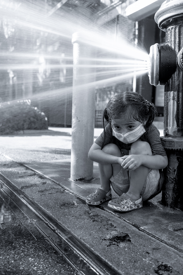 A girl sits crouched under a open fire hydrant.