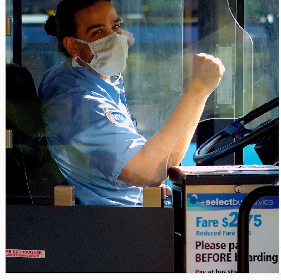 A bus driver wearing a mask sits at the wheel of a bus with his fist slightly raised.