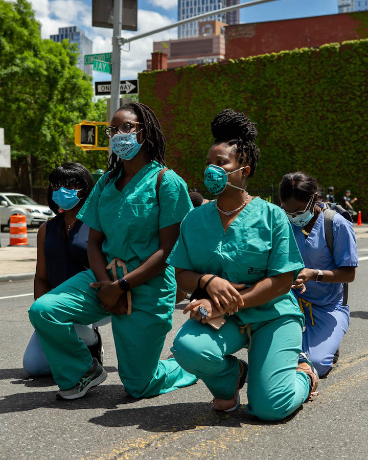 Four nurses in scrubs an masks kneel in a parking lot.