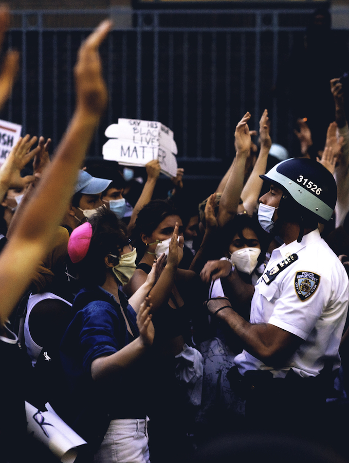 A group of protesters stand with their arms raised up, a cop in a mask and helmet stands facing them.