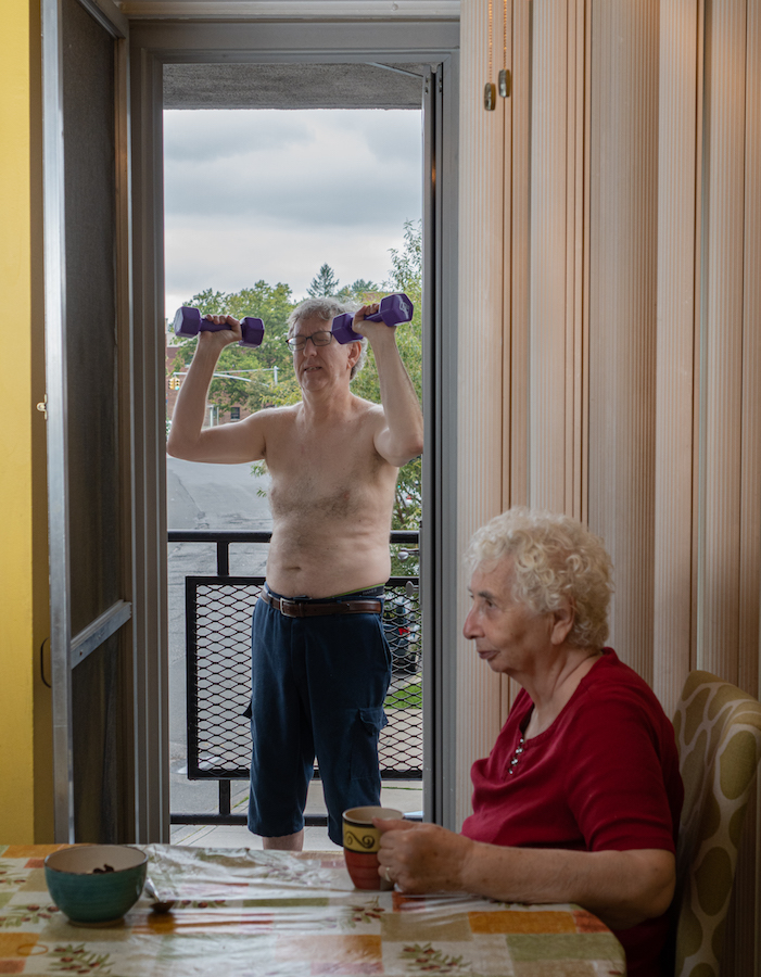 Um homem segurando pesos fica em uma porta em uma varanda Uma mulher está sentada dentro de casa lendo um jornal em uma mesa em primeiro plano.