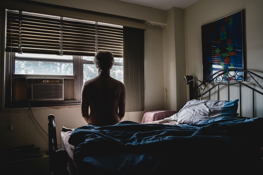 Un hombre se sienta en una cama sin camisa, frente a su ventana con una unidad de aire acondicionado instalada.