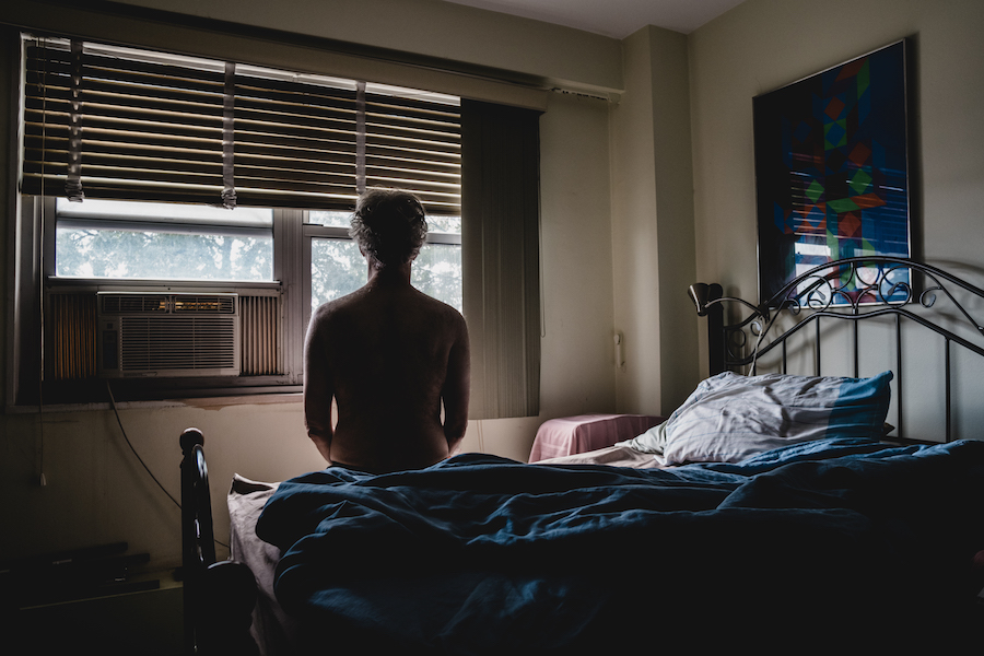 A man sits on a bed shirtless, facing his window with an AC unit installed.