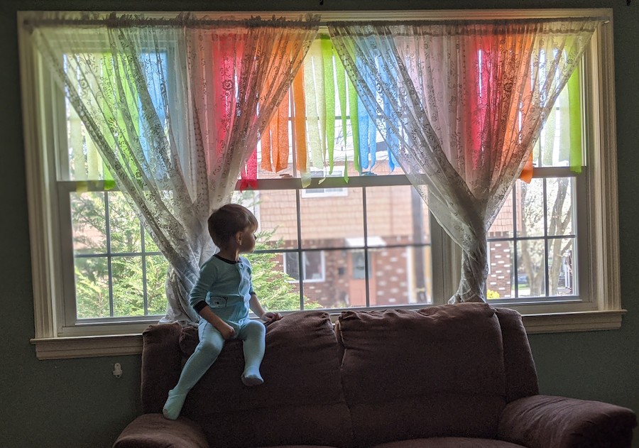 一个小男孩坐在沙发的靠背顶部，从窗帘和彩色的彩带覆盖的窗户向外看。