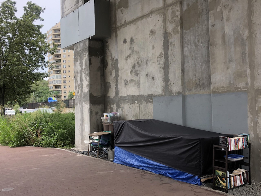 A long covered canvas structure, a metal bookshelf with many books, and a desk, chair, and other living supplies are carefully places along the side of a concrete building