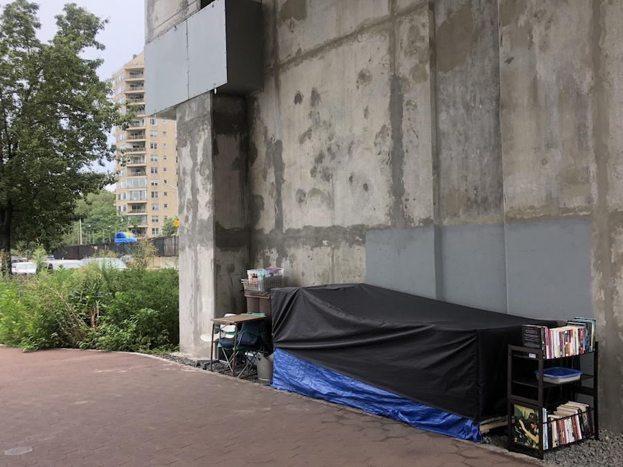 Uma longa estrutura de lona coberta, uma estante de metal com muitos livros e uma mesa, cadeira e outros suprimentos vivos são cuidadosamente colocados ao longo da lateral de um edifício de concreto