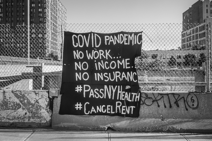 Uma fotografia em preto e branco de uma placa em uma cerca que diz "COVID PANDEMIC No work .... No Income ... No Insurance ... # PassNYHealth #CancelRent"