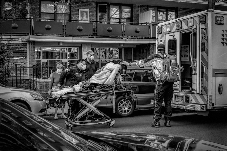 Une personne sur une civière est transportée dans une ambulance avec l'aide de trois travailleurs médicaux. Une quatrième personne se tient à proximité.
