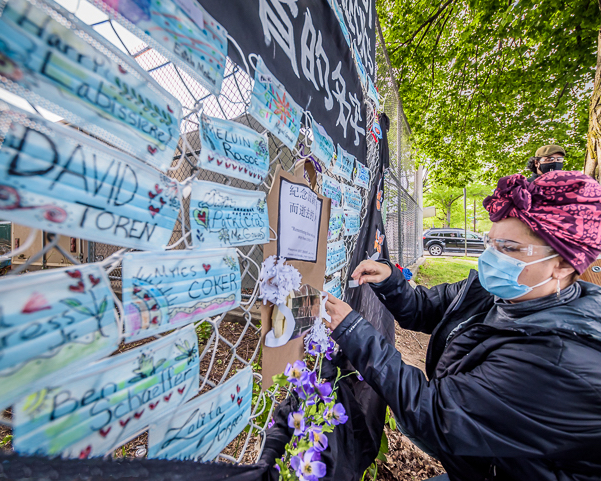 A woman places a mask with a name written on it on a fence covered in other blue face masks with names.