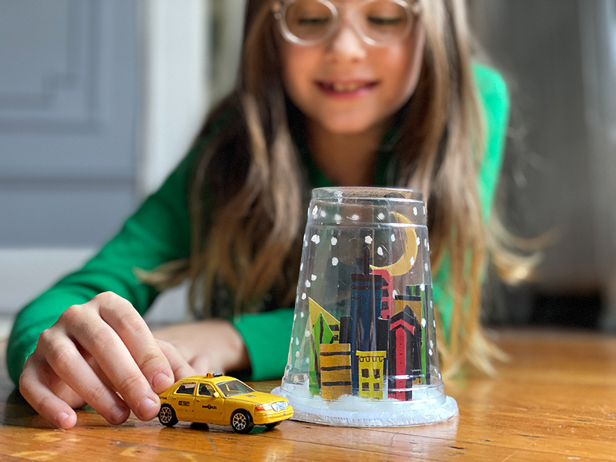 A young girl pushing a toy NYC taxi cab next to a snow globe made of a plastic cup with a hand-drawn city skyline  inside.