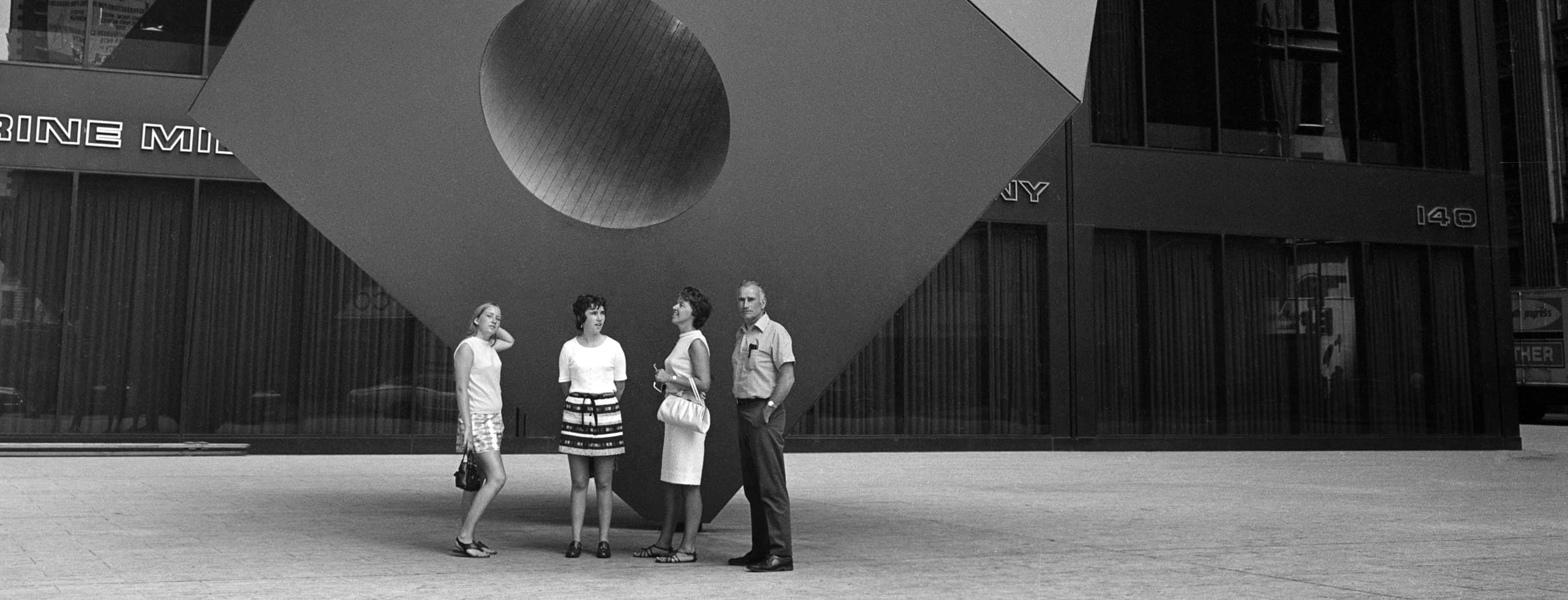 Four people stand on the sidewalk in front of a large statue of a cube with a hole going through the center of it