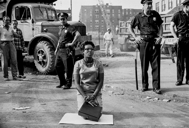 Downstate Medical Center建筑工地1963年CORE纠察队的一名抗议者跪在工人和警察的面前，阻止了建筑的进行。