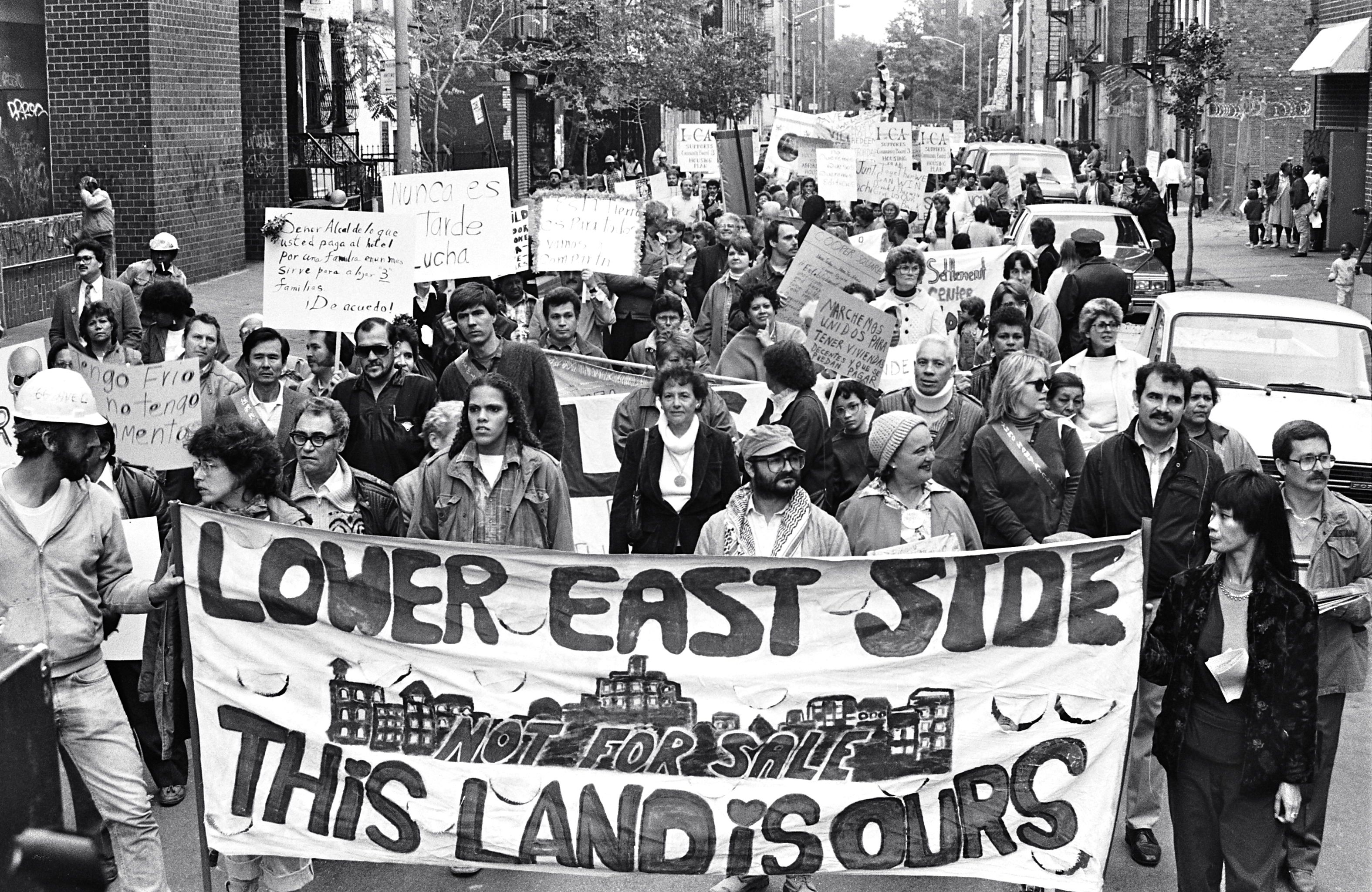 Une photo en noir et blanc d’un groupe d’adultes marchant dans une rue du Lower East Side. Les gens devant brandissent une banderole qui dit : « Lower East Side. Cette terre est à nous ».