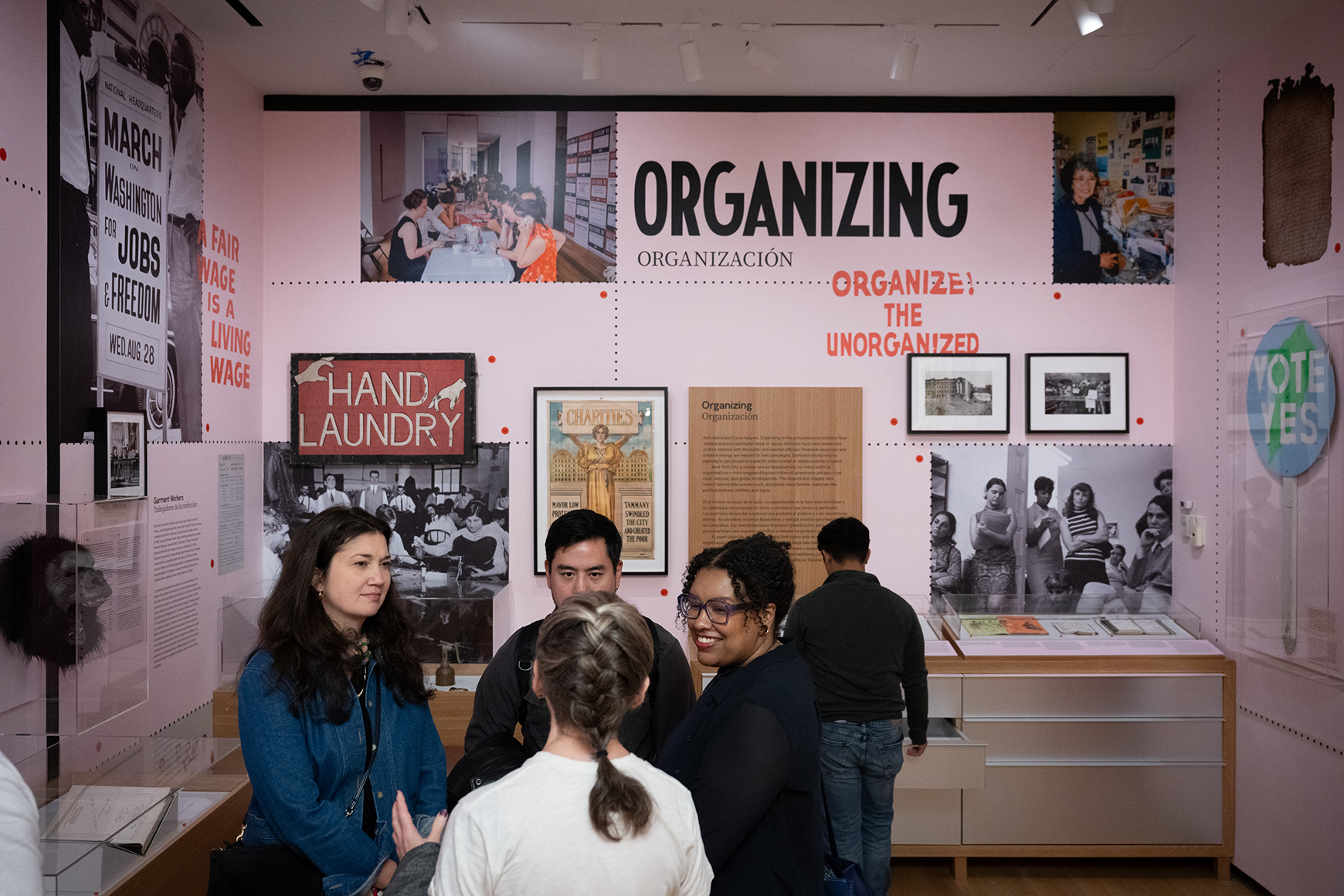 A crowd stands in a gallery with photographs, posters, and objects on pink walls