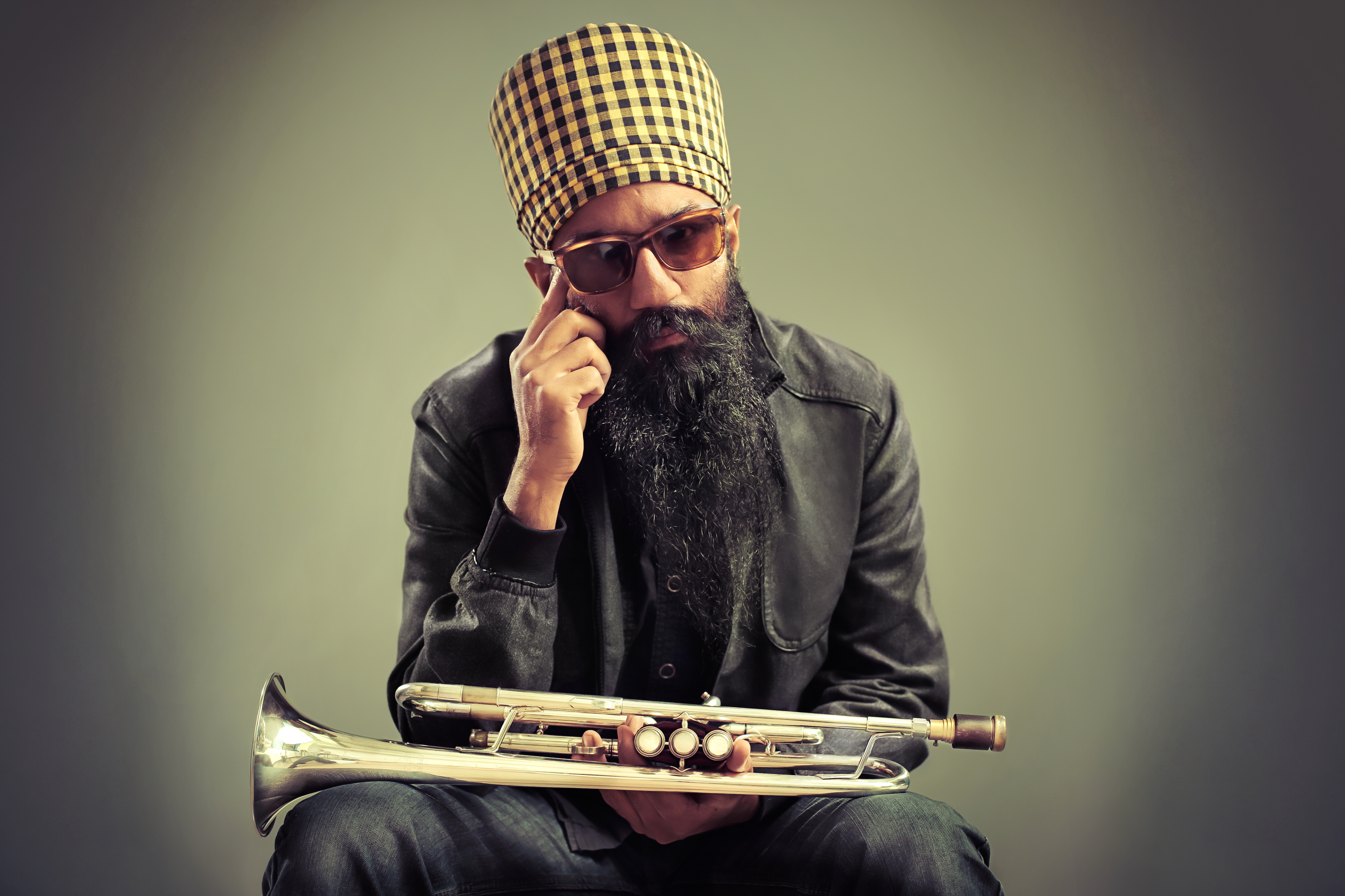 A man poses while wearing a checkered turban and holding a trumpet.  