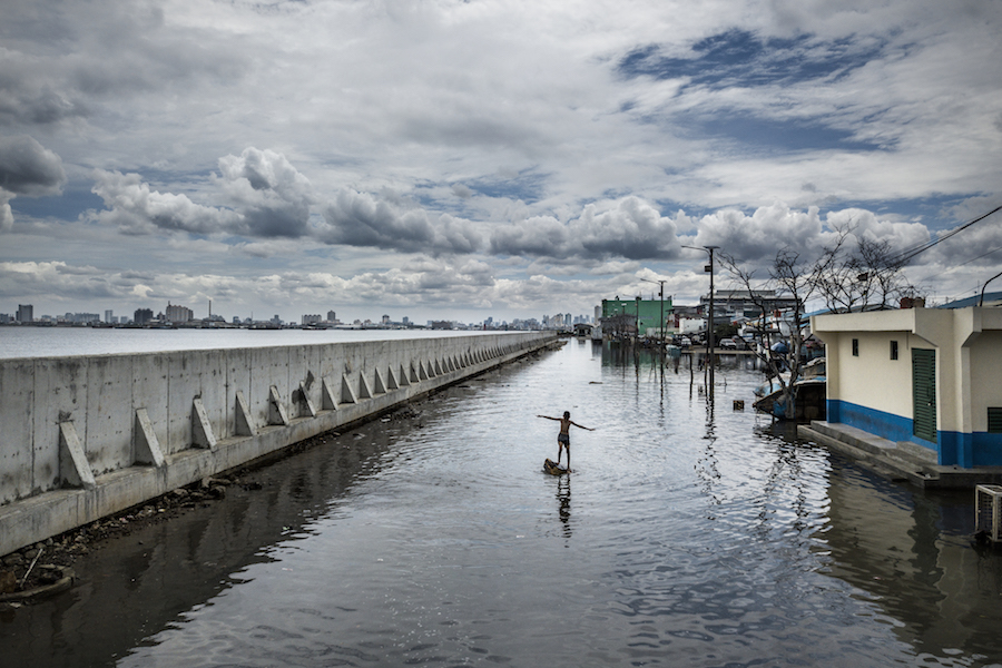 A boy stands on a box in a flooded section of Jakarta, between rows of houses and other buildings and a large sea wall. 