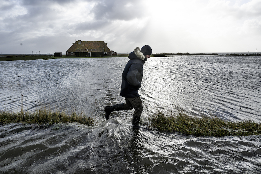 A man in a heavy coat runs across a flooded area following a thing stretch of grass. A small house is visible in the distance on another strip of land.  