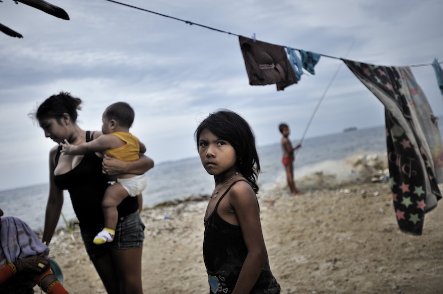 A mother, holding a baby, and a small child stand on a beach under a laundry line