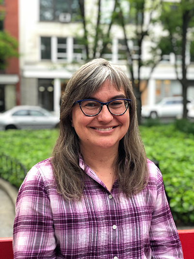 Headshot of a smiling woman wearing dark blue reading glasses and a plaid, purple shirt outdoors.