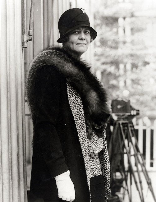 루스 베이커 프랫(Ruth Baker Pratt)이 야외 건물에 서 있는 사진. 그녀는 모직 모자, 장갑, 모피 스톨을 착용하고 있습니다.