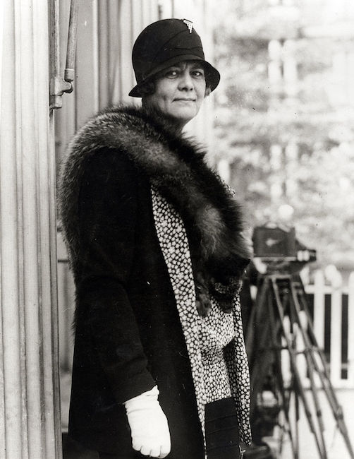 Fotografía de Ruth Baker Pratt de pie contra un edificio al aire libre. Lleva un gorro de lana, guantes y una estola de piel.