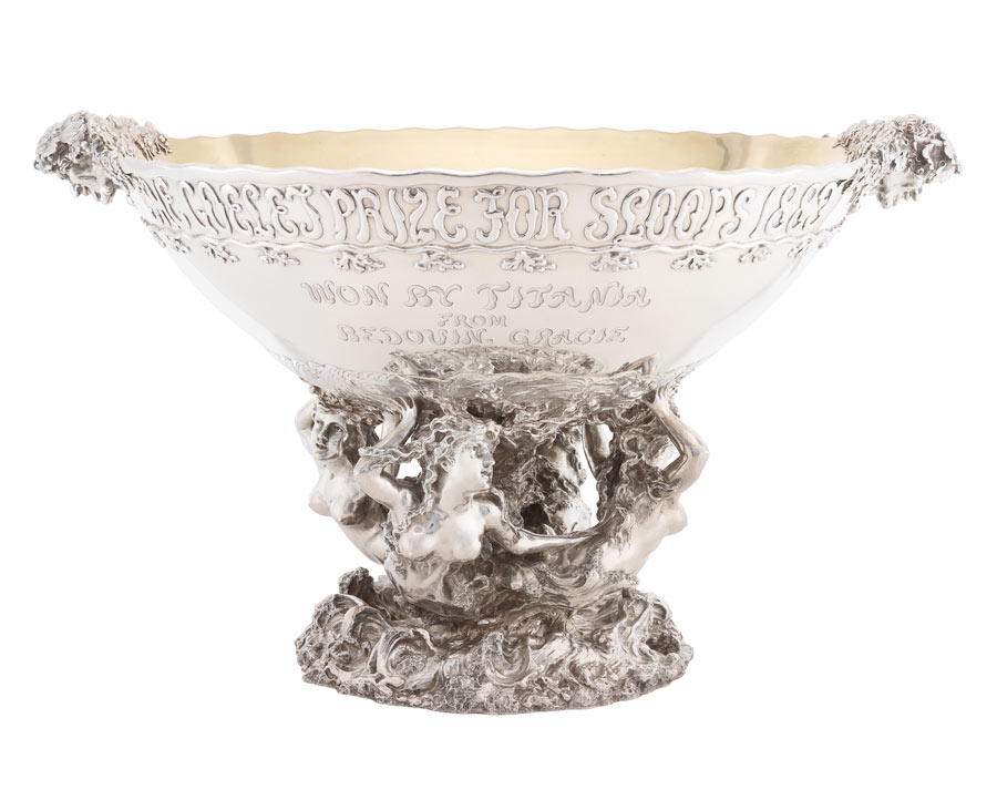 A large silver bowl held by statues of mermaids, with heads as handles and text on the bowl